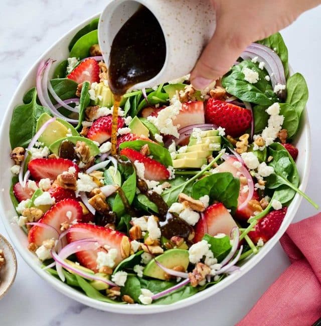 pouring Balsamic Poppy Seed dressing over spinach salad with strawberries and feta cheese