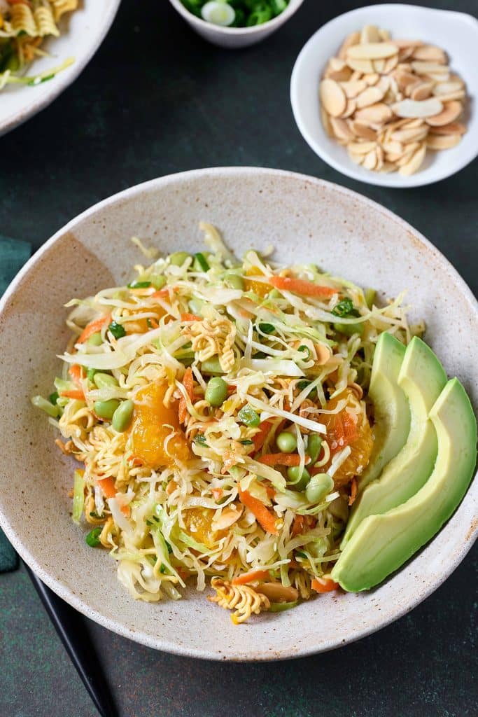 Top view of a bowl filled with cabbage slaw, mandarin oranges edamame and crispy ramen noodles tossed and served with slices of avocado.