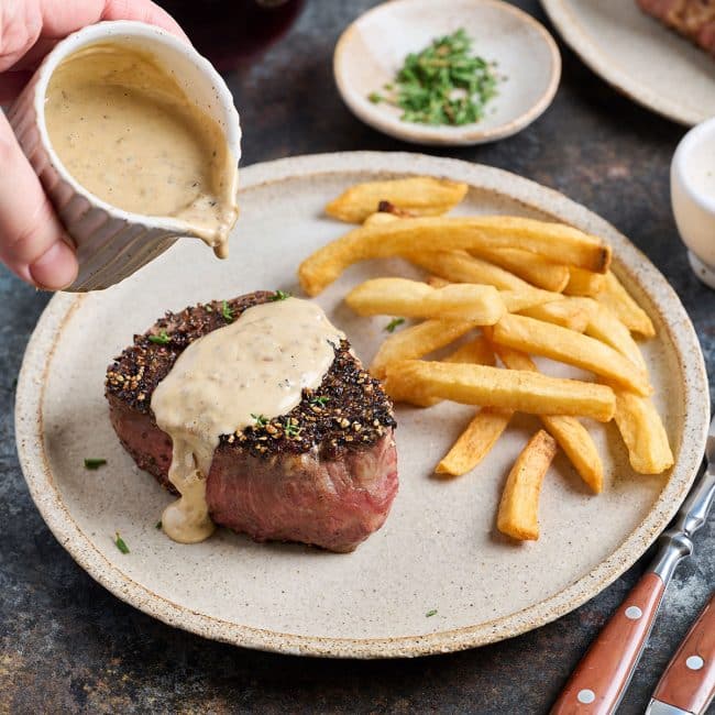 Pouring a silky and creamy sauce over a peppered steak.