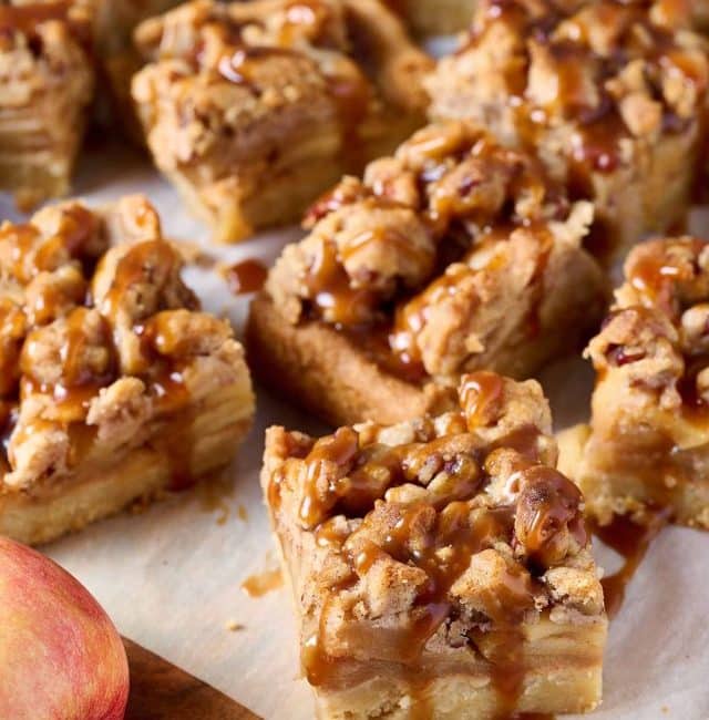 Apple pie bars with pecan crumble and drizzled with salted caramel sitting on pieces of parchment paper on top of a wooden board