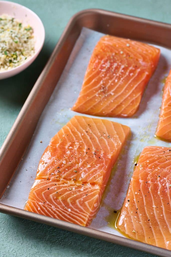 seasoned salmon filets on parchment paper line baking sheet with bowl of panko mixture next to it