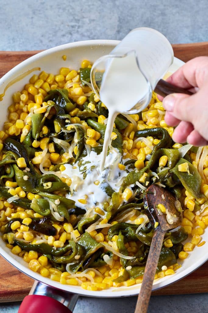 Pouring heavy cream into a skillet with roasted sliced poblano peppers, corn and sliced onions.