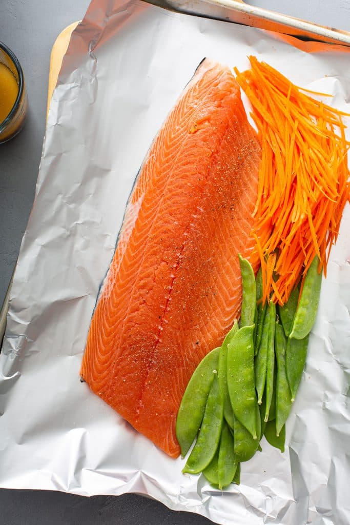 Large piece of salmon sitting on top of foil with carrots and peas placed on the side. 