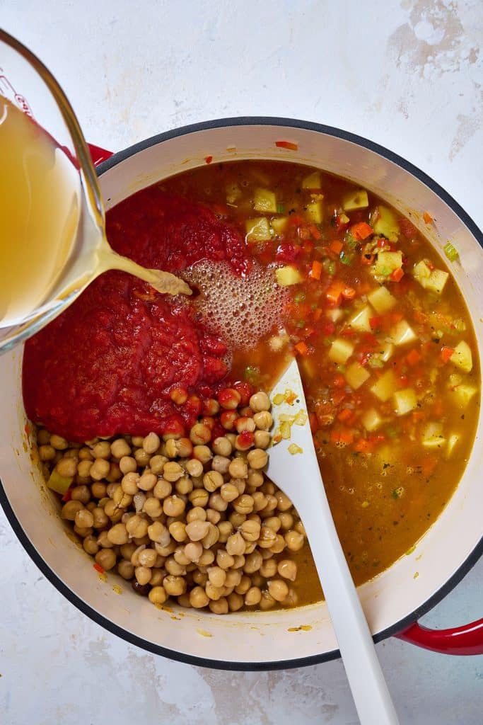 vegetable broth is being poured into a soup pot filled with vegetables, potatoes, garbanzo beans, and tomatoes.
