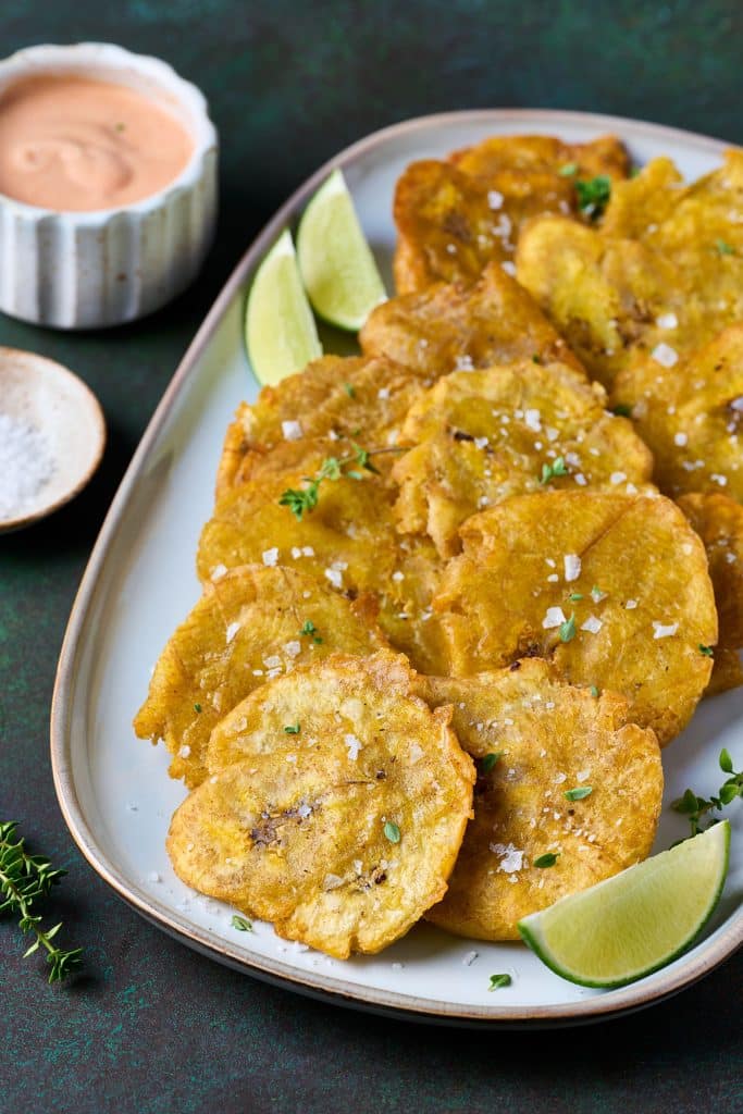 Fried green plantains (Tostones) laid out on a plate garnished with salt and lemon wedges and mayo ketchup on the side