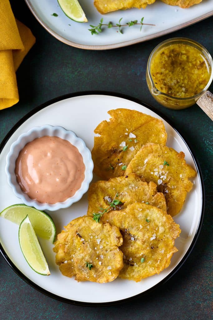 Patacones beautifully displayed on a plate. with lemon slices on the side and a bowl of kethcup mayo to be used for dipping.