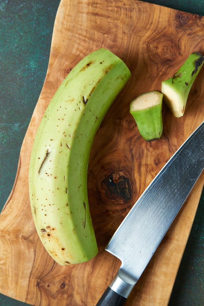 A green plantain on a cutting board with both ends chopped off with a knife.