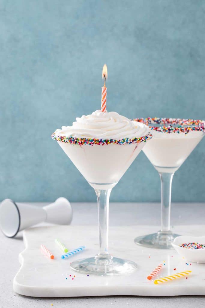 two martini glasses filled with birthday martini dipped in sprinkles and topped with piped whipped cream and a lit candle.