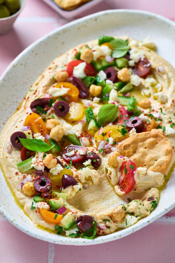 A platter of hummus topped with cherry tomatoes, cucumbers, olives, chickpeas, feta, basil, and spices, with a hand dipping a cracker into it.