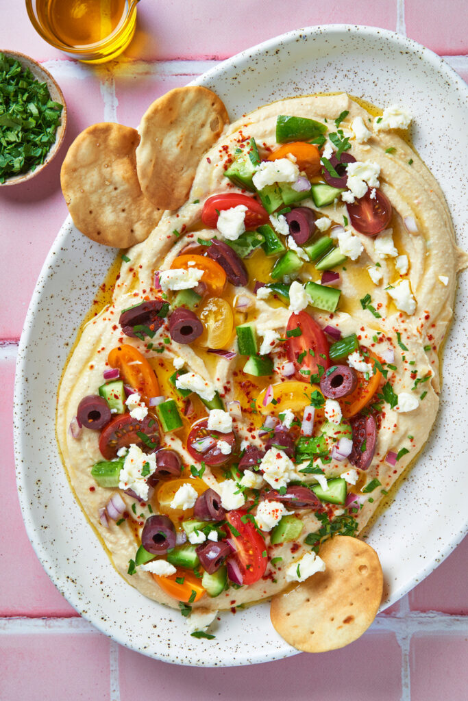 A platter of hummus topped with colorful cherry tomatoes, cucumbers, Kalamata olives, feta cheese, red onion, parsley, and a drizzle of olive oil, served with crispy pita rounds on the side.