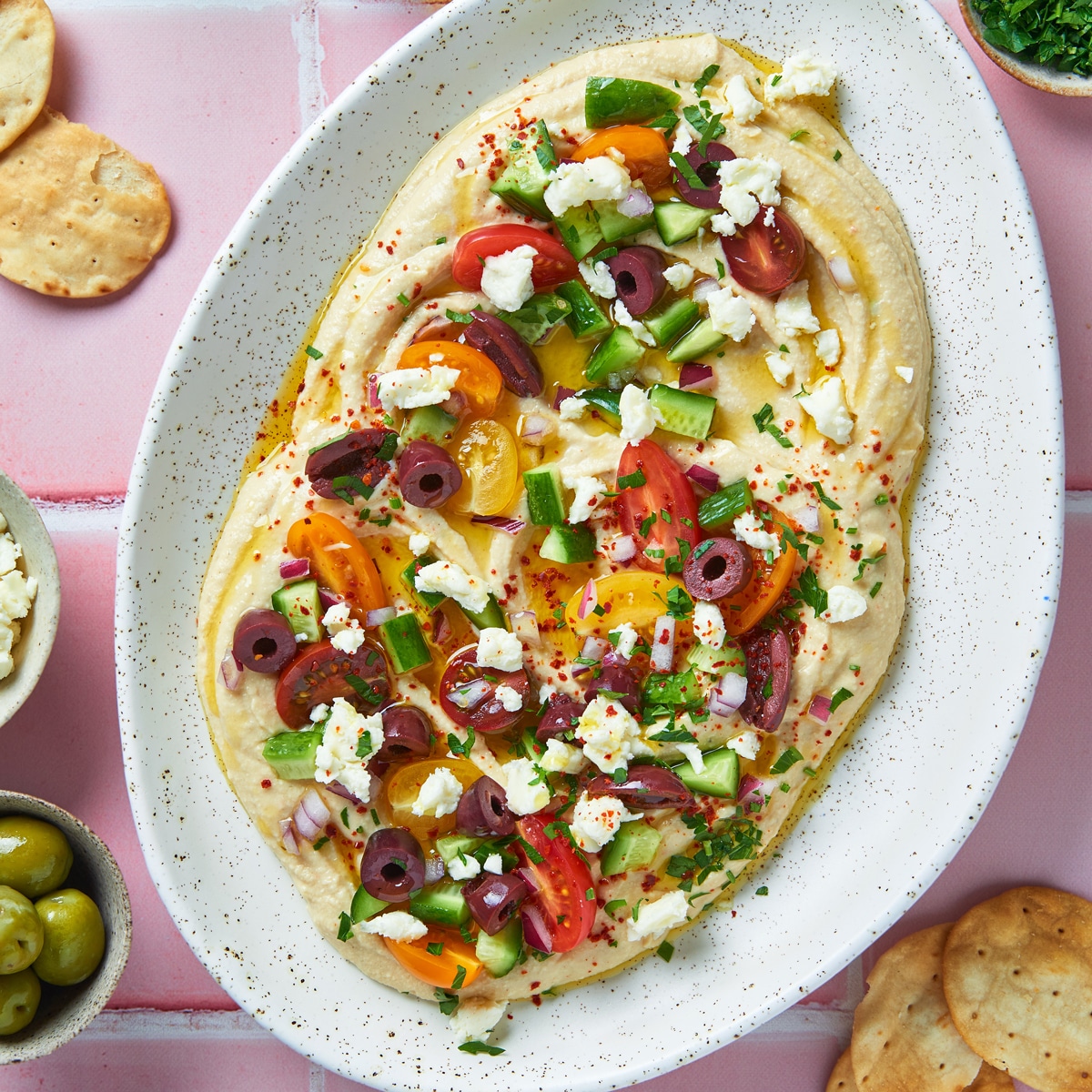 A platter of hummus topped with colorful cherry tomatoes, cucumbers, Kalamata olives, feta cheese, red onion, parsley, and a drizzle of olive oil, served with crackers on the side.