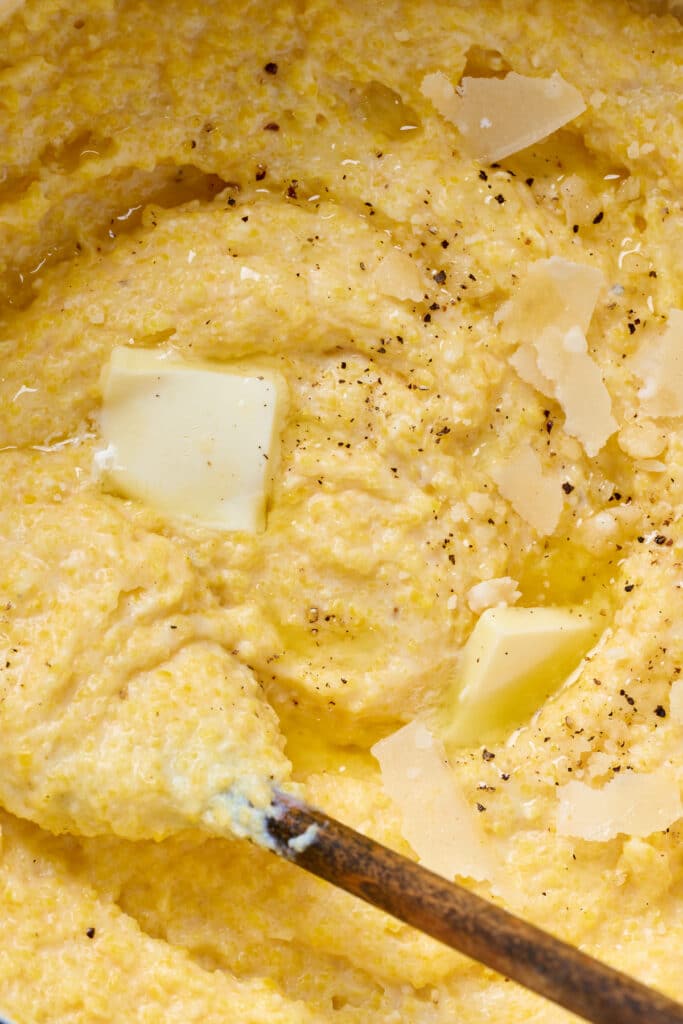 A wooden spoon stirring creamy polenta topped with melty butter and Parmesan cheese.
