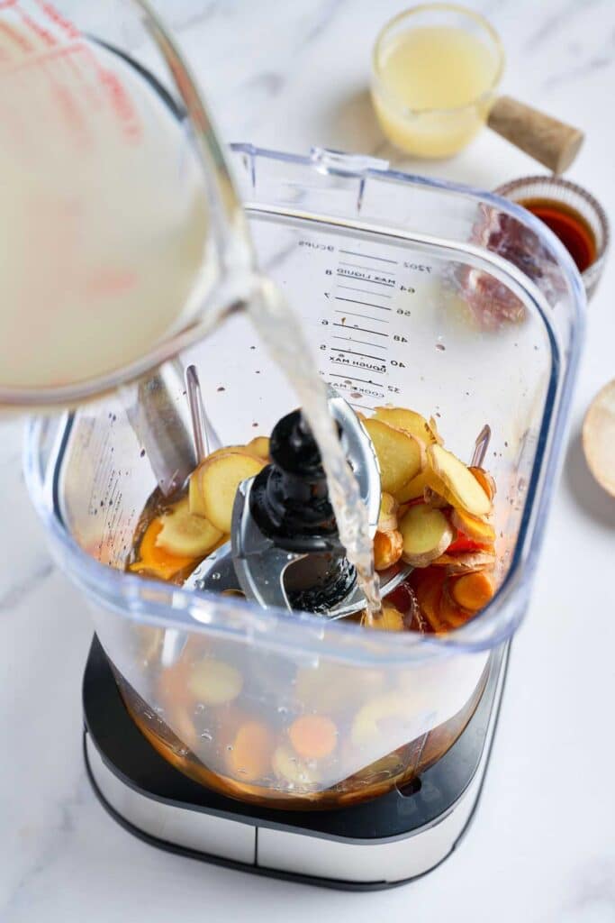 Coconut water is poured into a blender filled with sliced fresh turmeric and fresh ginger 