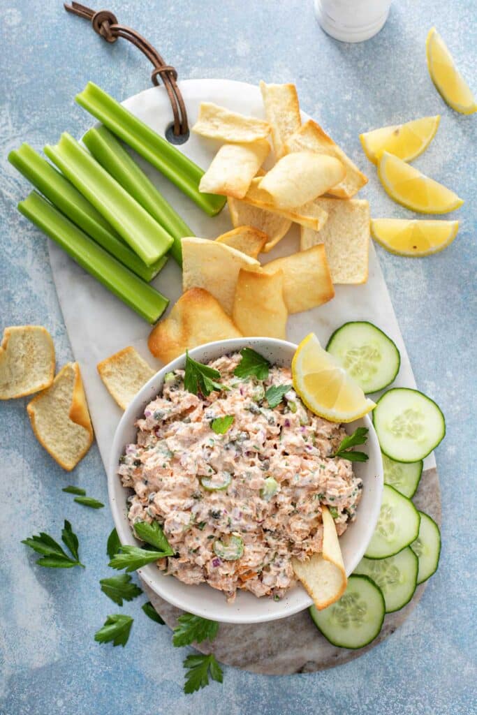 A bowl of creamy salmon salad on a marble board served with celery sticks, crackers and cucumber rounds.
