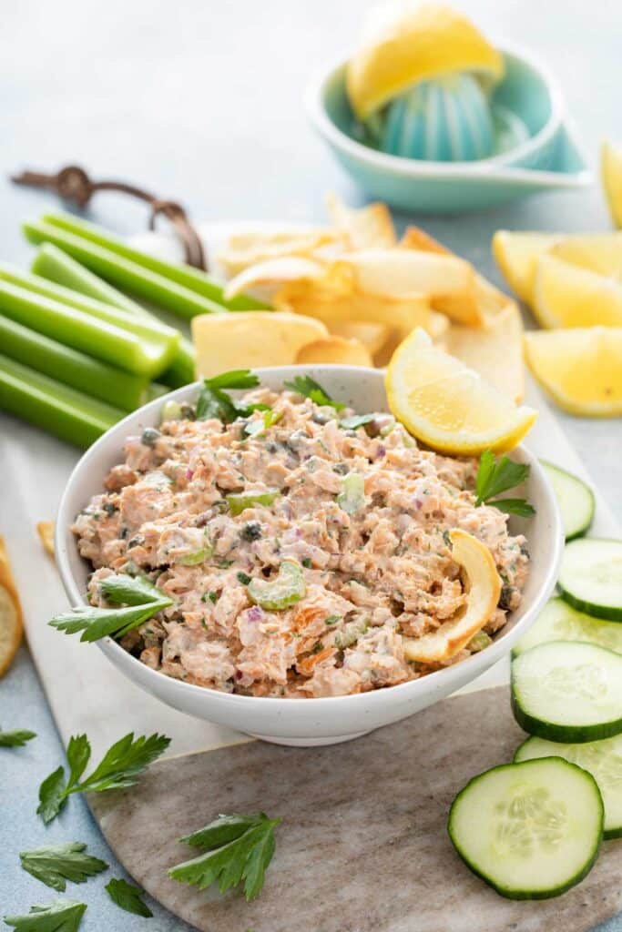 A bowl with creamy salmon salad with celery and onion, served with cucumber slices and garnished with lemon