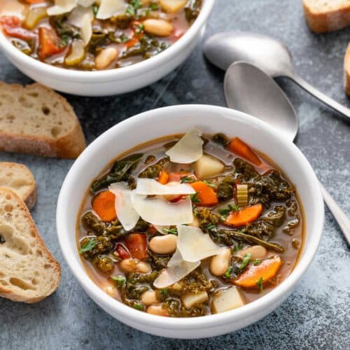 Two bowls of white bean soup with vegetables and greens topped with Parmesan shavings surrounded with crusty bread