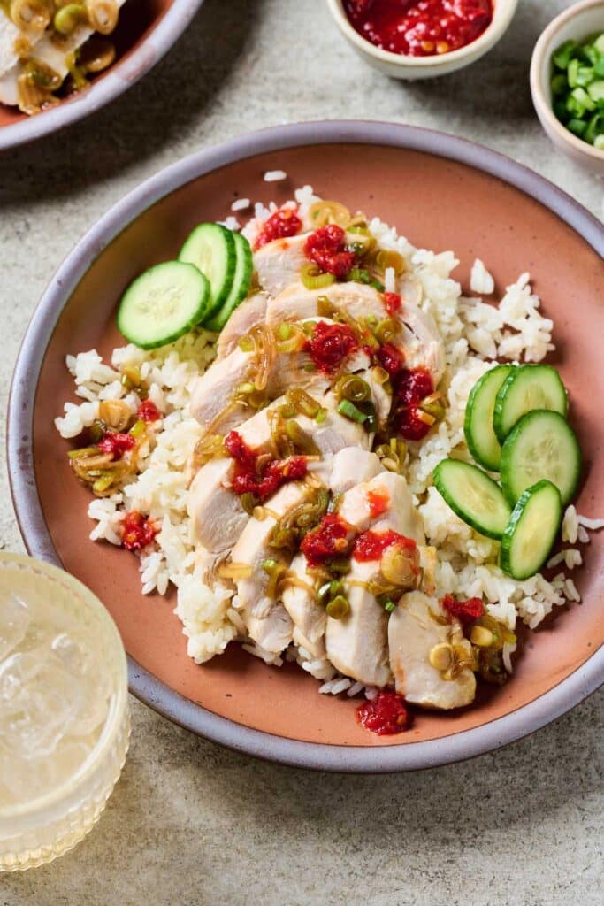 A plate of fluffy rice topped with poached and sliced Hainanese chicken topped with scallion oil and chili sauce and served with sliced cucumbers.