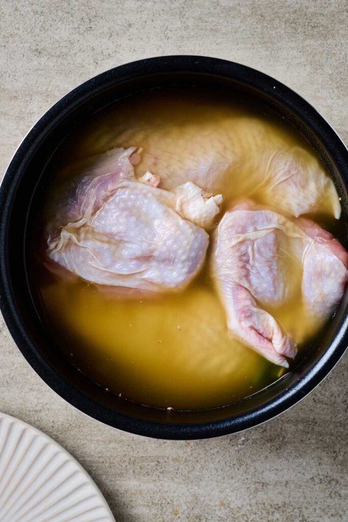 Pieces of skin-on chicken pieces submerge in broth in a rice cooker