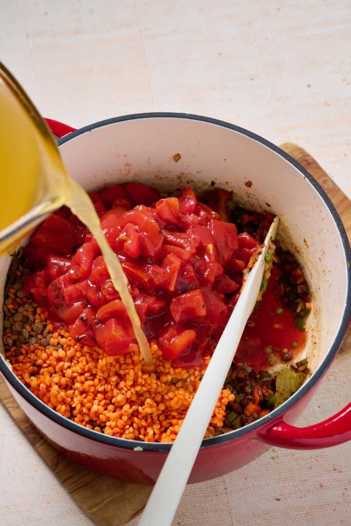 Adding broth into a pot with sautéed veggies, red and brown lentils, and canned roasted diced tomatoes.