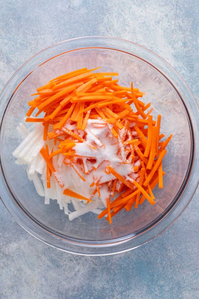 Daikon radish and carrot sticks in a glass bowl sprinkled with sugar and salt
