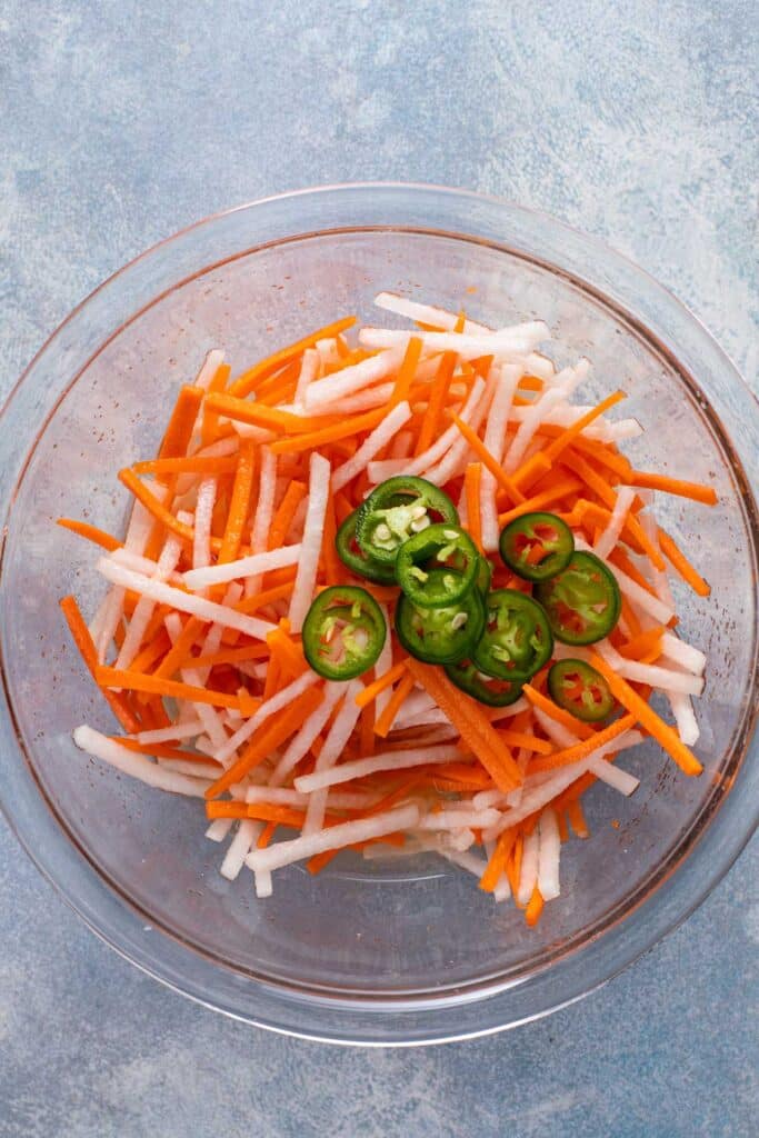 Daikon radish sticks and carrot sticks in a glass bowl topped with sliced jalapeños.