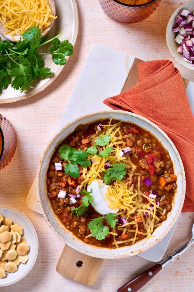A bowl of lentil chili with veggies topped with shredded cheddar cheese, cilantro, chopped red onion and sour cream