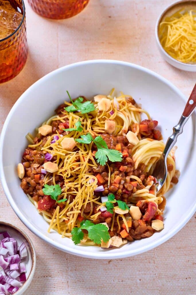 A bowl of lentil's chili over spaghetti and garnish with oyster crackers, red onion, cilantro and shredded cheese.