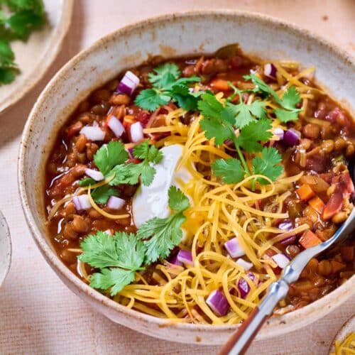 A bowl of lentil chili topped with chopped red onions, cilantro and shredded cheese