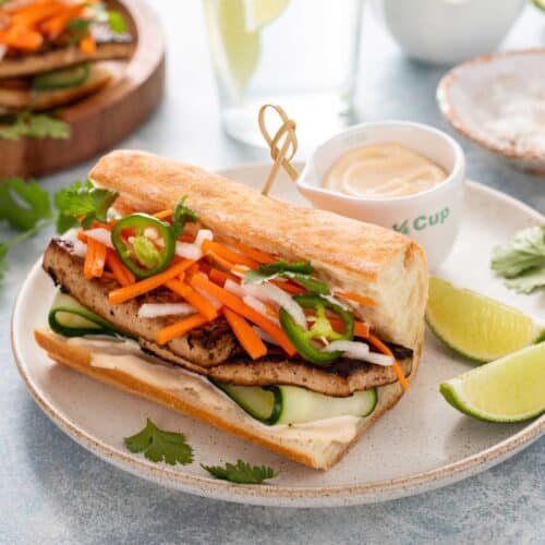 A tofu sandwich on baguette bread topped with pickled veggies and cilantro and garnished with lime wedges