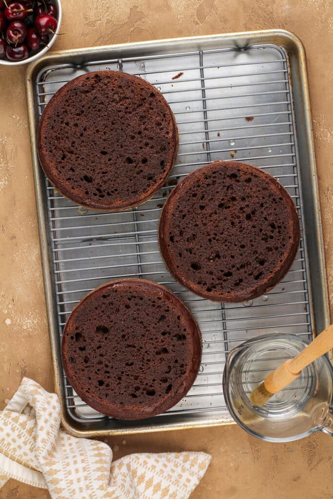 three layers of chocolate cake on a cooling rack next to a bowl with syrup and a pastry brush