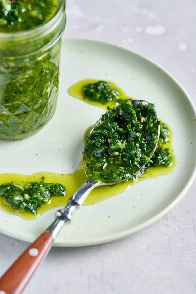 A spoon spilling an Argentinian condiment over a plate, next to a jar filled with the herby condiment