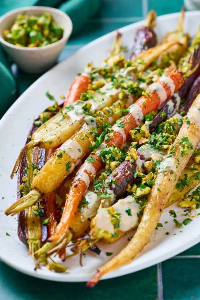 A platter with roasted multi-colored carrots topped with fresh herbs and chopped pistachios and a drizzle of tahini sauce.