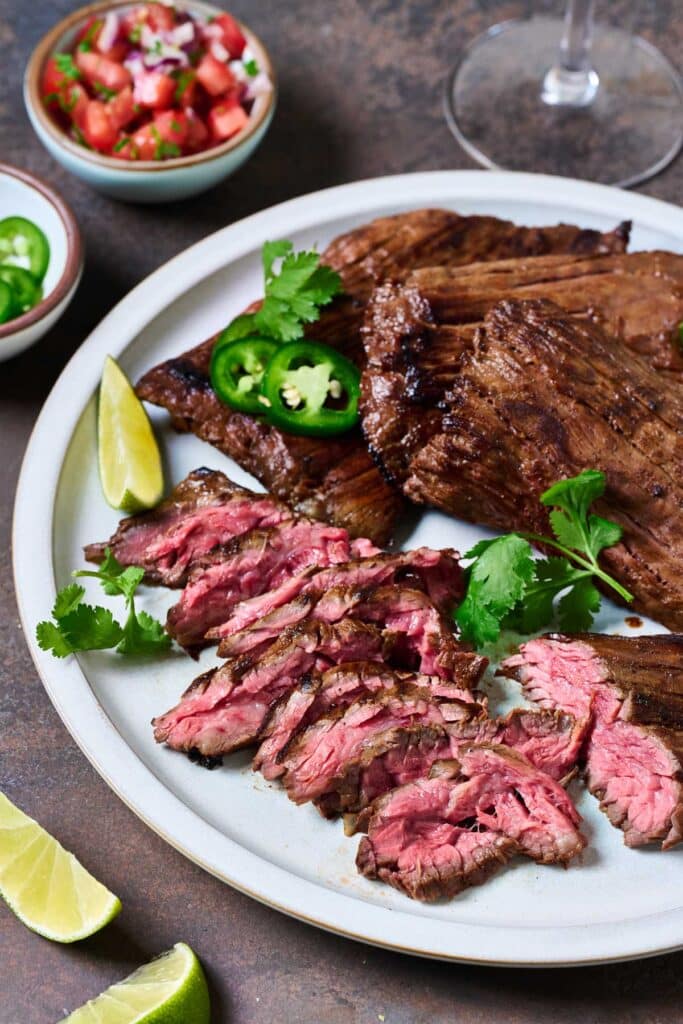 Grilled skirt steak on a plate with some of the steak sliced into bite size pieces, garnished with limes, cilantro and jalapeño slices