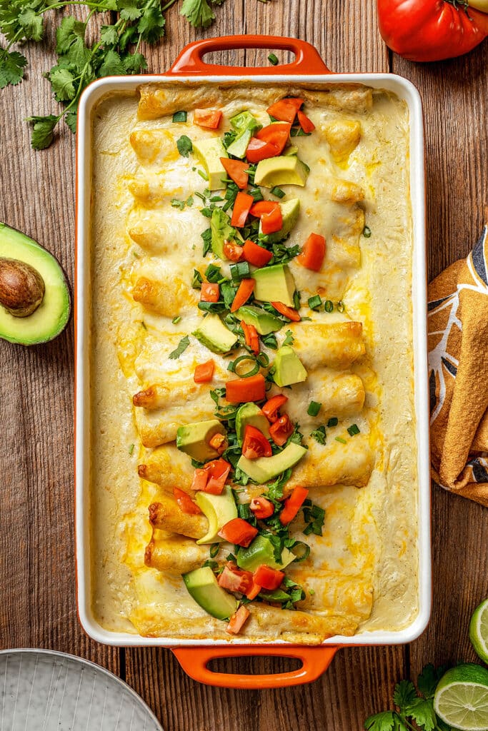 a casserole baking dish with rolled enchiladas topped in a creamy and cheesy sauce topped with chopped tomatoes, avocado and cilantro