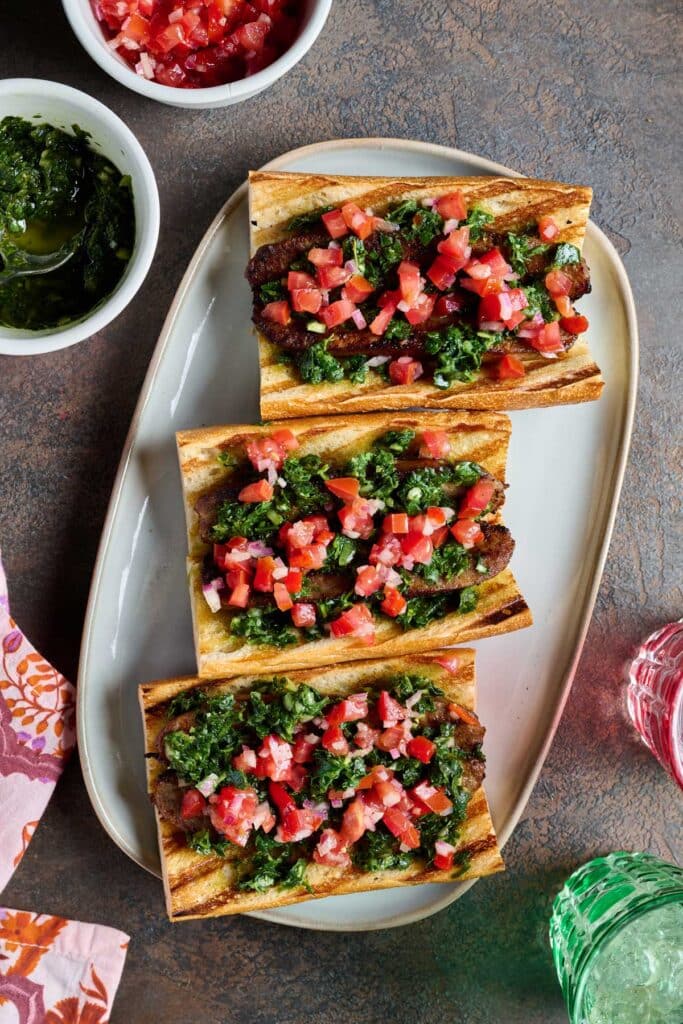 Three sausage sandwiches topped with herb chimichurri and chopped tomatoes and onions