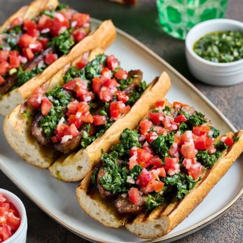 Three sausage sandwiches on crusty bread topped with chimichurri sauce and tomato and onion salsa