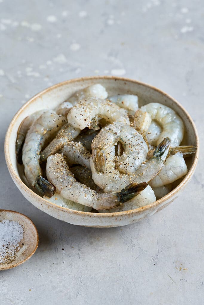 Raw shrimp with tails on in a bowl seasoned with salt and pepper