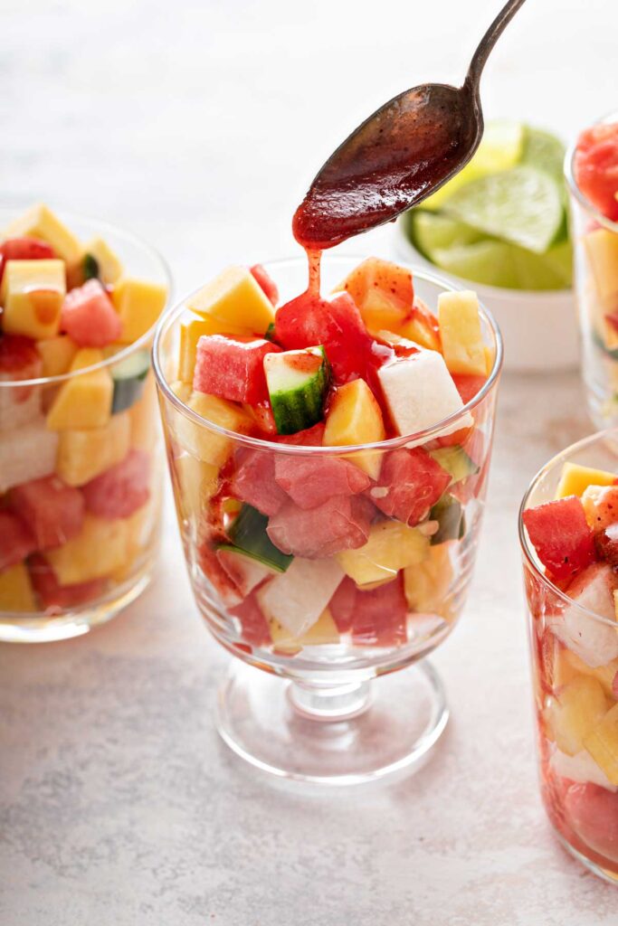A spoon drizzling chamoy sauce over fresh fruit pieces in a glass cup