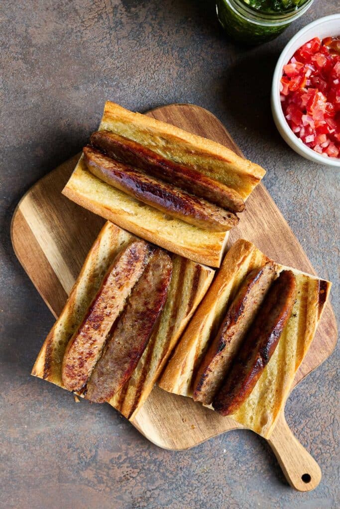 Grilled bread topped with golden brown sausages cut lengthwise on a wooden cutting board