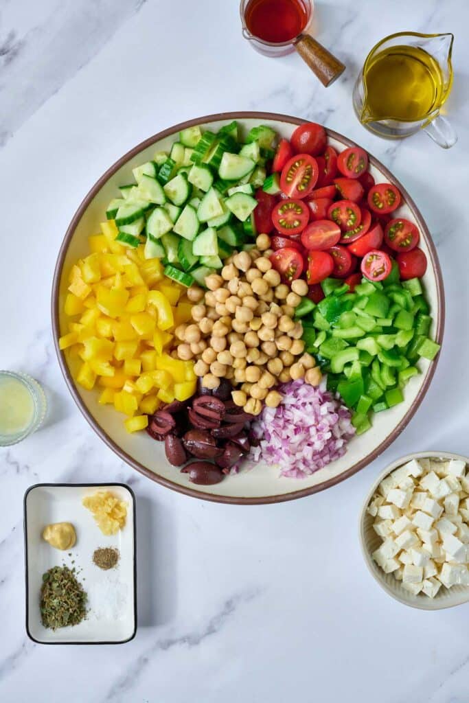 a bowl with cut up cherry tomatoes, chopped cucumbers, chickpeas, bell peppers, red onion and Kalamata olives next to a small bowl of feta cheese