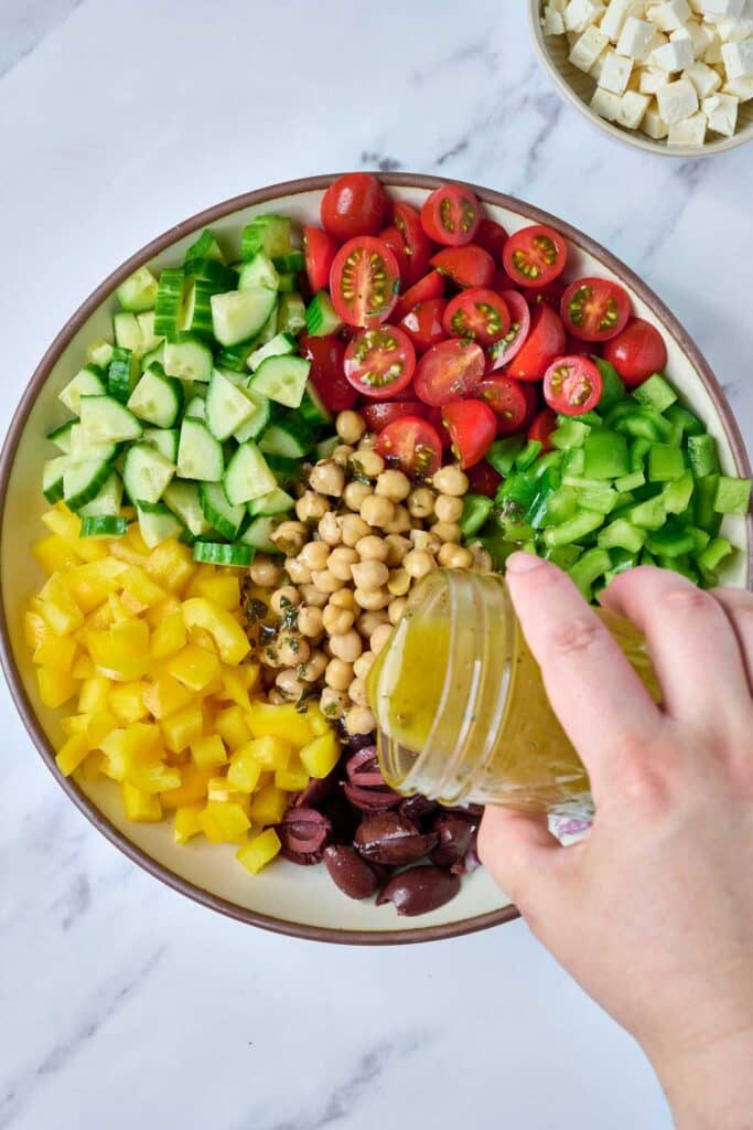 Pouring salad dressing over fresh chopped veggies, chickpeas and olives