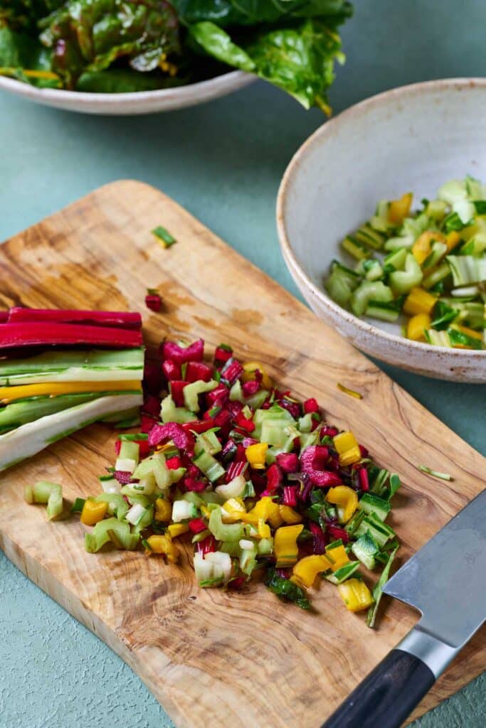 Cutting rainbow Swiss chard stems into small pieces with a knife on a wooden cutting board.