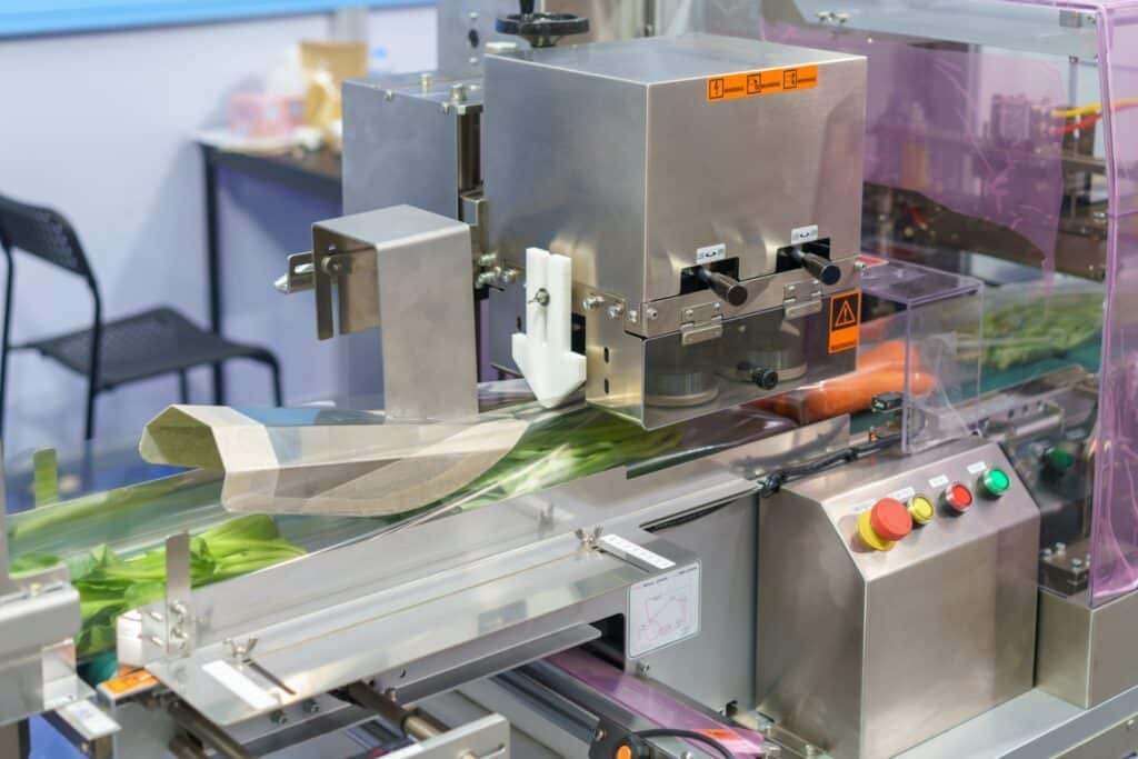 A stainless steel automated packaging machine wraps vegetables in plastic film on a conveyor belt in a commercial food processing facility.