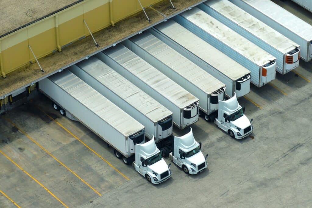 An aerial view of multiple white semi-trucks with refrigerated trailers backed into numbered loading dock bays at a large distribution warehouse.