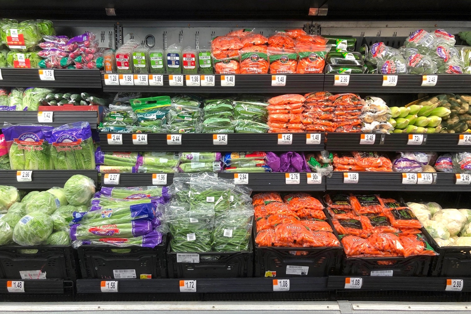 Refrigerated grocery store shelves stocked with rows of pre-packaged vegetables including bagged carrots, mini cucumbers, celery, and fresh herbs with price tags visible.
