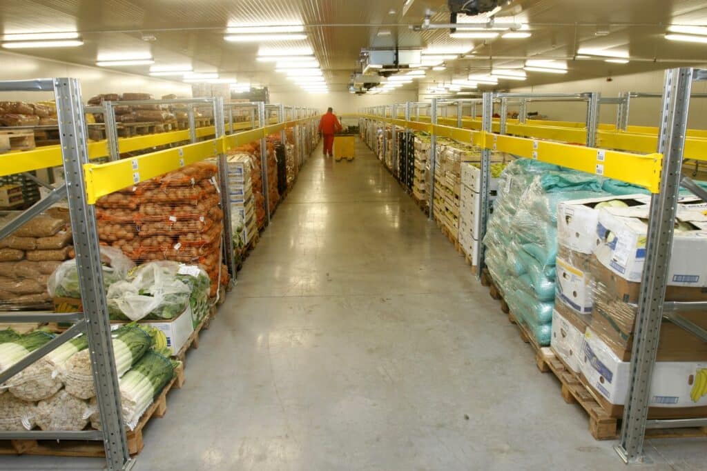 A long, brightly lit cold storage warehouse with pallets of vegetables including leeks, potatoes, and boxed produce stacked on metal shelving on both sides, with a worker visible in the distance.