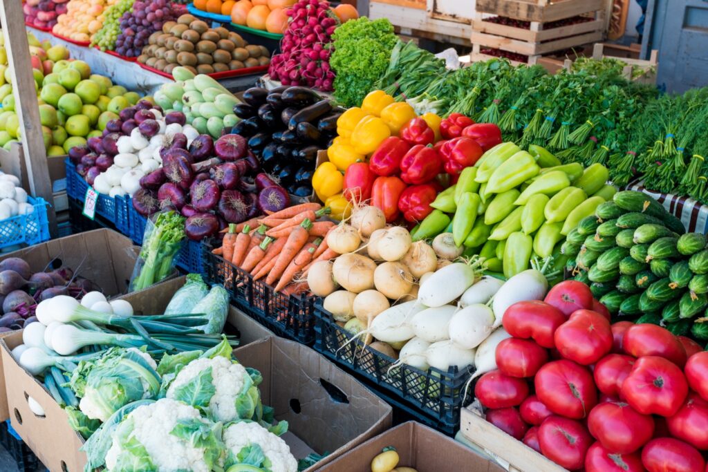 A vibrant outdoor market stall overflowing with an abundant variety of fresh vegetables and fruits, including bell peppers, eggplant, carrots, cauliflower, tomatoes, and radishes arranged in colorful rows.