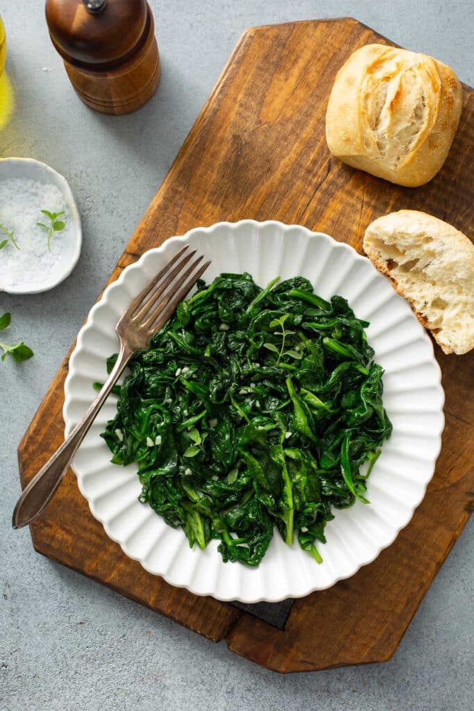 A white plate with bright green sautéed spinach on a cutting board. Crusty bread is also shown on the photo