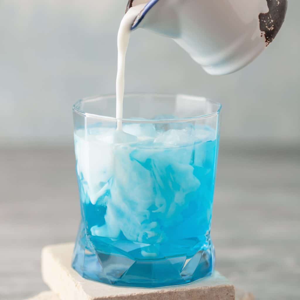 Cream being poured from a small pitcher into a faceted rocks glass filled with bright blue blueberry soda and ice, creating white swirling clouds through the drink.