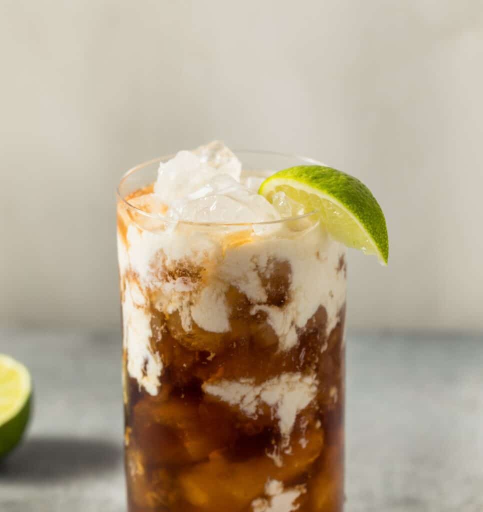 A tall glass of classic dirty soda with cola and cream swirled through crushed ice, garnished with a lime wedge, with a halved lime on the surface beside it.